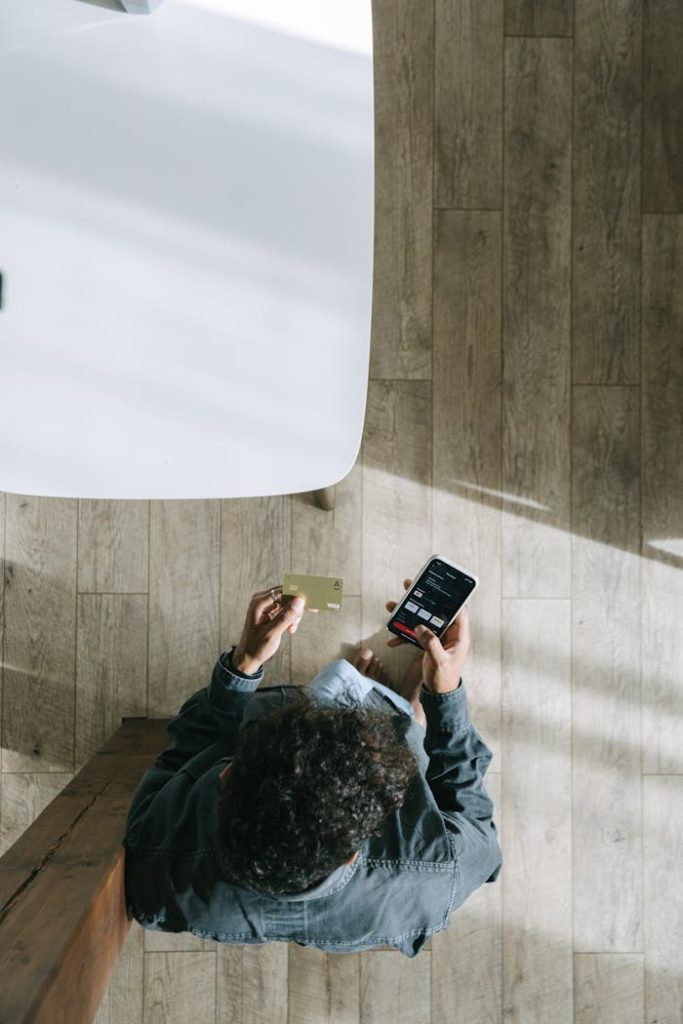 a-man-holding-a-card-while-using-his-smartphone-7620908 Man using smartphone and credit card for online shopping indoors, top view.