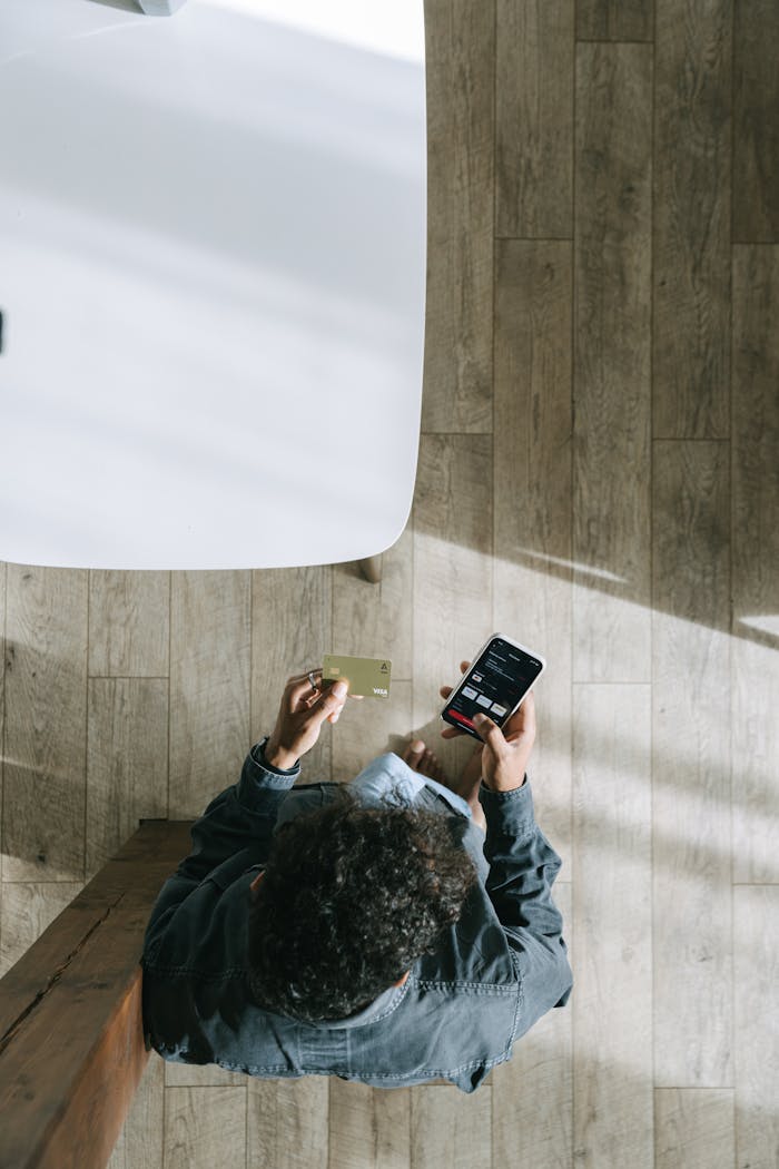 Man using smartphone and credit card for online shopping indoors, top view.